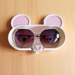 Pode incluir: Um suporte branco em forma de coelho para &oacute;culos de sol com detalhes de orelhas rosa e um nariz cinza. O suporte cont&eacute;m &oacute;culos de sol com arma&ccedil;&atilde;o prateada e lentes degrad&ecirc;. O suporte est&aacute; sobre uma superf&iacute;cie de madeira clara.