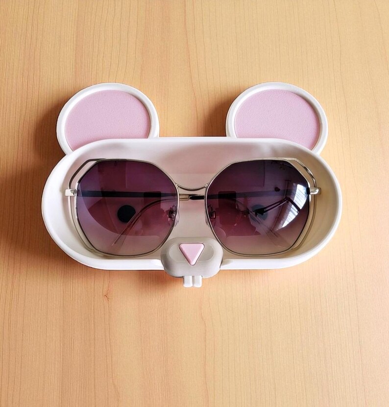 Pode incluir: Um suporte branco em forma de coelho para &oacute;culos de sol com detalhes de orelhas rosa e um nariz cinza. O suporte cont&eacute;m &oacute;culos de sol com arma&ccedil;&atilde;o prateada e lentes degrad&ecirc;. O suporte est&aacute; sobre uma superf&iacute;cie de madeira clara.