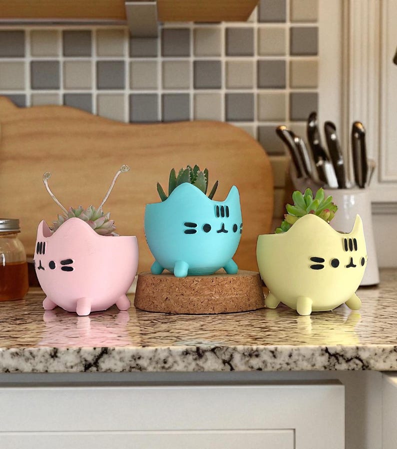 May include: Three cat-shaped succulent planters in pastel colors: pink, blue, and yellow. Each planter has a black face design and holds a small succulent plant. The planters are displayed on a countertop.