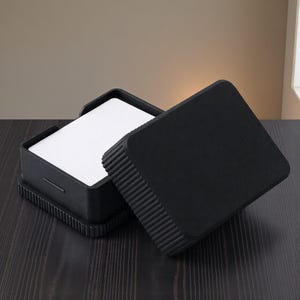 May include: A black square box with a textured lid, open to reveal a stack of white cards. The box has a ribbed design around the base and lid. The box is sitting on a dark wooden surface.