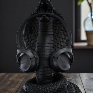 Op de afbeelding: Een zwarte koptelefoonstandaard in de vorm van een cobra met een zwarte JBL koptelefoon. De standaard is gedetailleerd, met de kop van de slang open om de koptelefoon vast te houden. De standaard staat op een houten oppervlak.