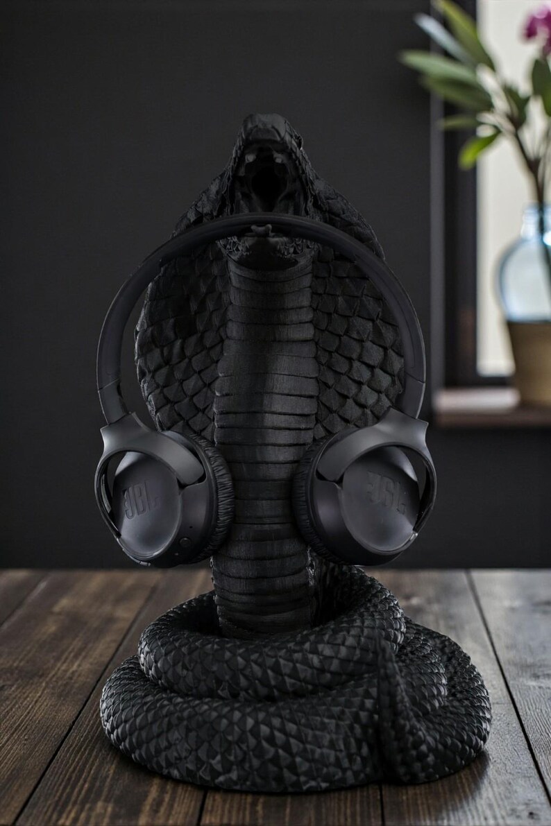 Op de afbeelding: Een zwarte koptelefoonstandaard in de vorm van een cobra met een zwarte JBL koptelefoon. De standaard is gedetailleerd, met de kop van de slang open om de koptelefoon vast te houden. De standaard staat op een houten oppervlak.