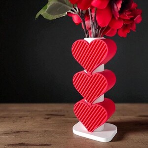 K&ouml;nnte beinhalten: Eine rote herzf&ouml;rmige Vase mit wei&szlig;em Sockel und Stiel, die rote Blumen h&auml;lt. Die Vase hat drei gestapelte Herzen mit diagonalen Linien. Der Hintergrund ist dunkel und die Vase steht auf einer Holzoberfl&auml;che.