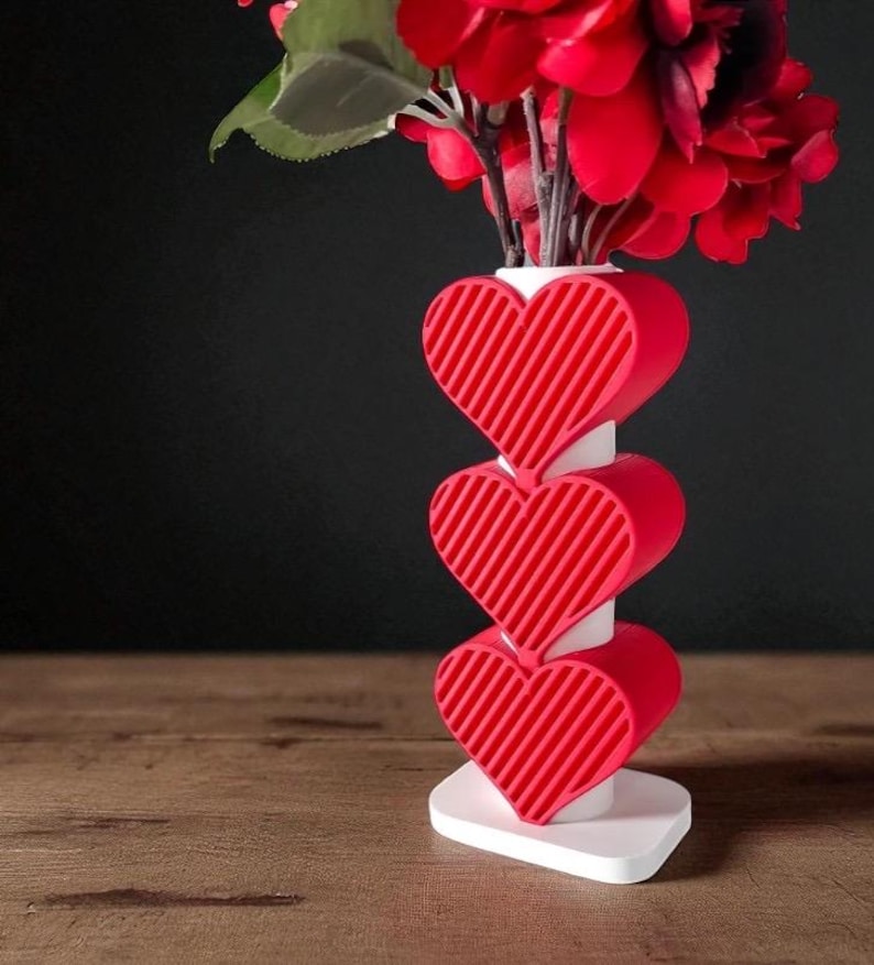 K&ouml;nnte beinhalten: Eine rote herzf&ouml;rmige Vase mit wei&szlig;em Sockel und Stiel, die rote Blumen h&auml;lt. Die Vase hat drei gestapelte Herzen mit diagonalen Linien. Der Hintergrund ist dunkel und die Vase steht auf einer Holzoberfl&auml;che.
