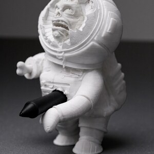 Pu&ograve; includere: Una statuetta di astronauta bianca stampata in 3D con un casco danneggiato che rivela una faccia mostruosa. Un oggetto cilindrico nero &egrave; inserito nel petto dell'astronauta. La statuetta &egrave; su una superficie grigia.