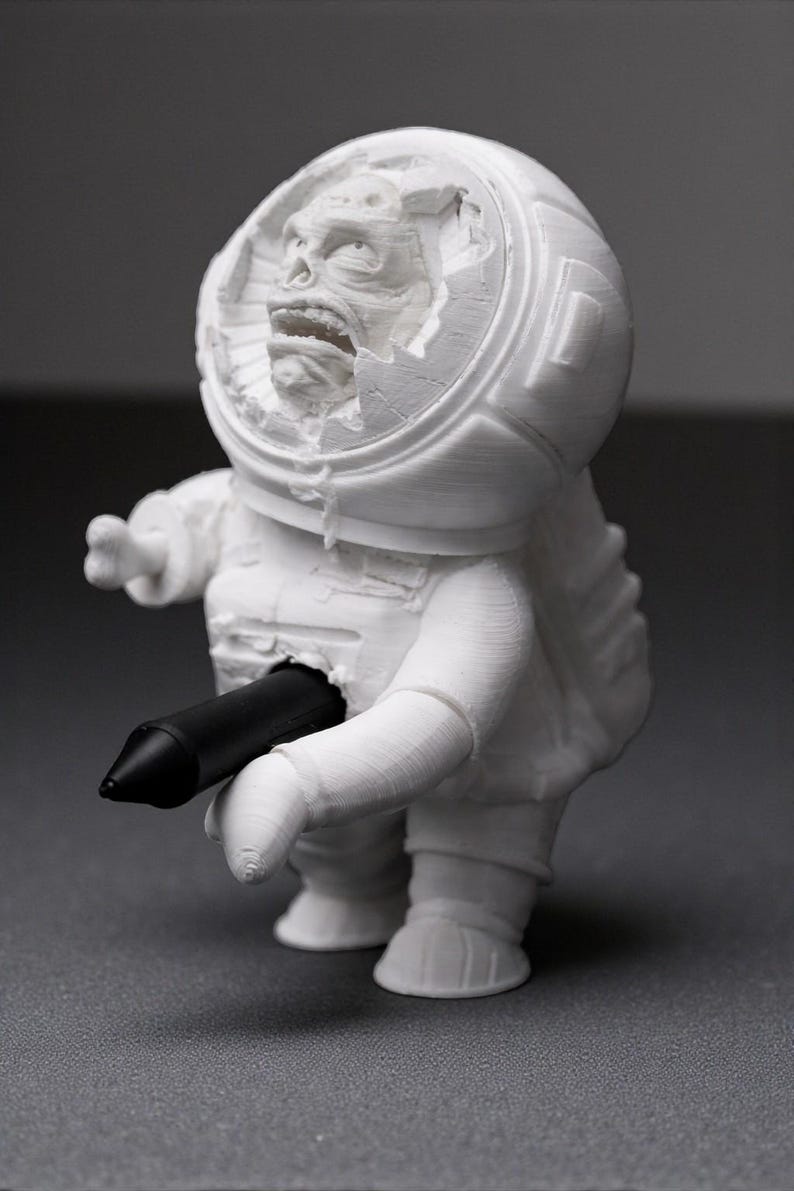 Pu&ograve; includere: Una statuetta di astronauta bianca stampata in 3D con un casco danneggiato che rivela una faccia mostruosa. Un oggetto cilindrico nero &egrave; inserito nel petto dell'astronauta. La statuetta &egrave; su una superficie grigia.