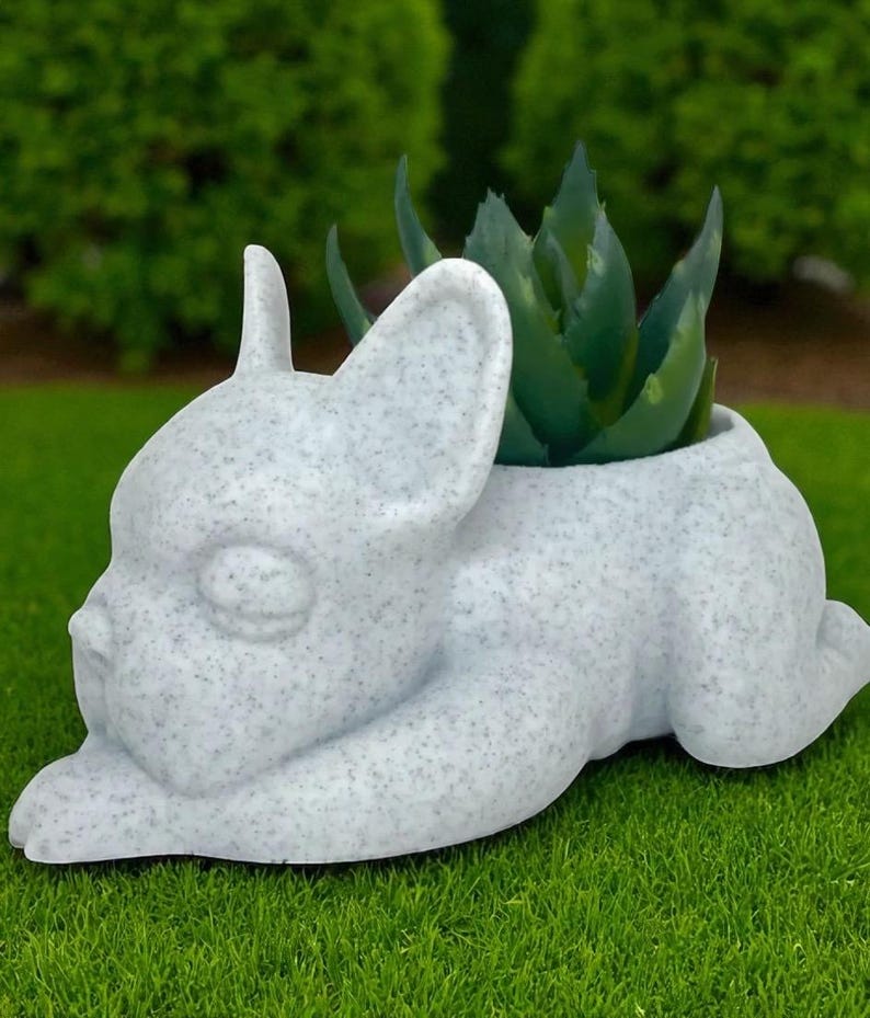 May include: A speckled white ceramic planter shaped like a French Bulldog, with a green succulent plant growing from its head. The dog is lying down on green grass, with a blurred green background.