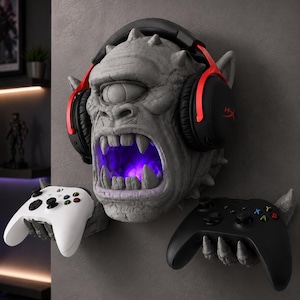 May include: A gray, stone-textured cyclops wall mount featuring a single eye and an open mouth with a purple internal glow. The creature has two hands extending outward to hold a white game controller on the left and a black game controller on the right, with black and red gaming headphones resting on its head.
