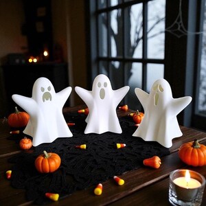 May include: Three white ghost figurines with outstretched arms are arranged on a table, surrounded by miniature pumpkins and candy corn. A lit candle in a glass holder adds to the Halloween theme. The ghosts have different facial expressions.