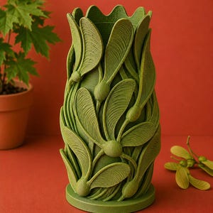 以下が含まれることがあります： カエデの種子の詳細なレリーフデザインが施された緑色の花瓶。花瓶は丸いベースと波状のトップエッジを持っています。背景は無地の赤色です。