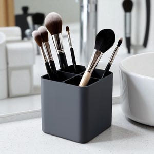 Peut inclure: Un porte-pinceaux de maquillage carré gris foncé avec six compartiments, rempli de divers pinceaux de maquillage. Les pinceaux ont des manches noirs et des poils doux dans les tons marron et blanc. Le support est posé sur un comptoir blanc.