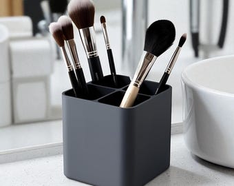 May include: A dark gray square makeup brush holder with six compartments, filled with various makeup brushes. The brushes have black handles and soft bristles in shades of brown and white. The holder sits on a white countertop.