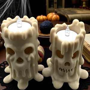 May include: Two off-white Halloween skull-shaped candles with dripping wax details. Each candle has a flickering LED light. The candles are on a black lace mat, with pumpkins and a dark background.