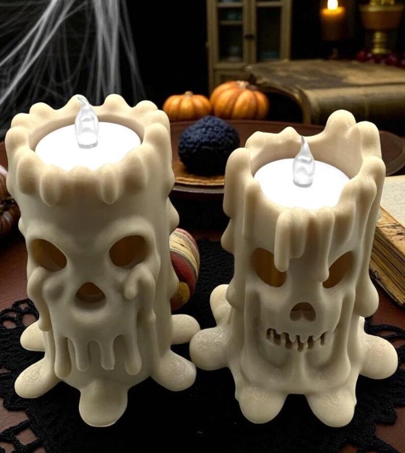 May include: Two off-white Halloween skull-shaped candles with dripping wax details. Each candle has a flickering LED light. The candles are on a black lace mat, with pumpkins and a dark background.