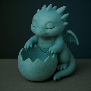Puede incluir: Figura de drag&oacute;n turquesa con los ojos cerrados, sosteniendo una c&aacute;scara de huevo agrietada. El drag&oacute;n tiene peque&ntilde;as alas y detalles puntiagudos en la espalda. La c&aacute;scara de huevo forma un peque&ntilde;o cuenco. Un art&iacute;culo decorativo caprichoso.