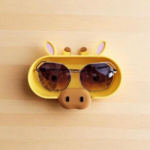 Puede incluir: Un soporte amarillo para gafas de sol con forma de cara de jirafa, con gafas de sol tintadas en marr&oacute;n en su interior. El soporte tiene orejas rosas, cuernos marrones y una nariz marr&oacute;n. Las gafas de sol tienen montura dorada.