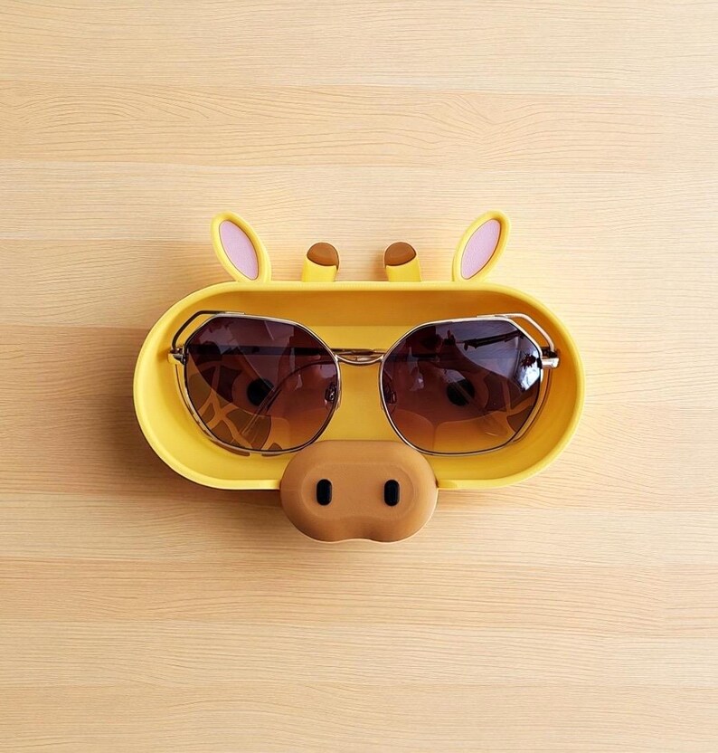 Puede incluir: Un soporte amarillo para gafas de sol con forma de cara de jirafa, con gafas de sol tintadas en marr&oacute;n en su interior. El soporte tiene orejas rosas, cuernos marrones y una nariz marr&oacute;n. Las gafas de sol tienen montura dorada.