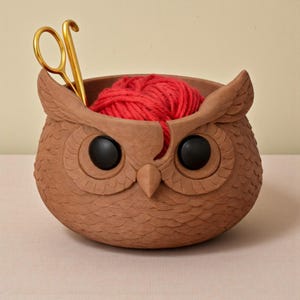 May include: A brown owl-shaped yarn bowl with black eyes and a beak, holding a ball of red yarn and gold scissors. The bowl has a textured surface and is designed to hold yarn for knitting or crocheting projects.