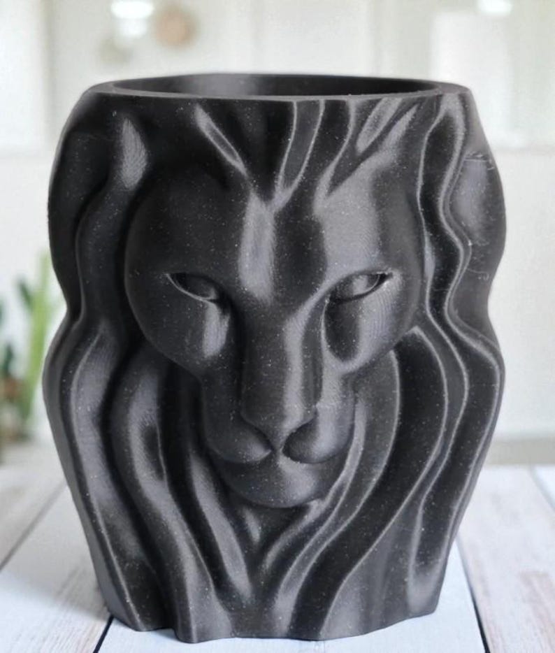 May include: A black, 3D-printed planter shaped like a lion's head. The planter features detailed facial features, including eyes, a nose, and a mane. The planter is designed to hold small plants or flowers and is approximately 15 cm tall.