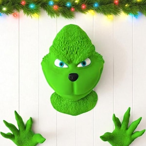 Pode incluir: Um conjunto de decoração de Natal com o tema Grinch verde. O conjunto inclui um rosto do Grinch com olhos azuis e nariz preto, e dois recortes de mãos verdes. Uma grinalda com luzes coloridas no topo.
