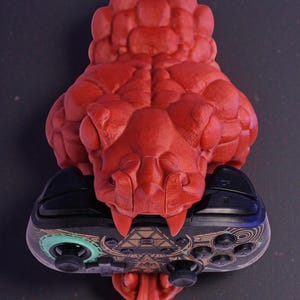 May include: A red 3D-printed snake head holds a black and patterned video game controller. The snake's head is detailed with scales and fangs, and its tongue extends downward. The controller has a geometric design.