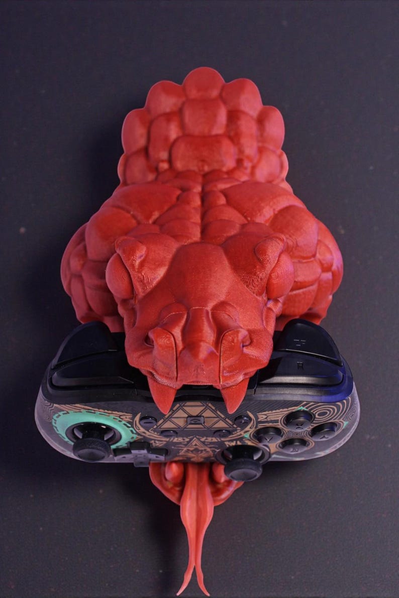 May include: A red 3D-printed snake head holds a black and patterned video game controller. The snake's head is detailed with scales and fangs, and its tongue extends downward. The controller has a geometric design.