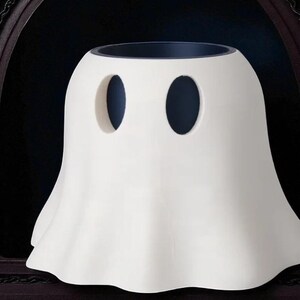 May include: A white ceramic ghost-shaped container with two oval eye-holes. The container has a wide, ruffled base and a dark interior. The design is simple and whimsical, perfect for Halloween or spooky decor.