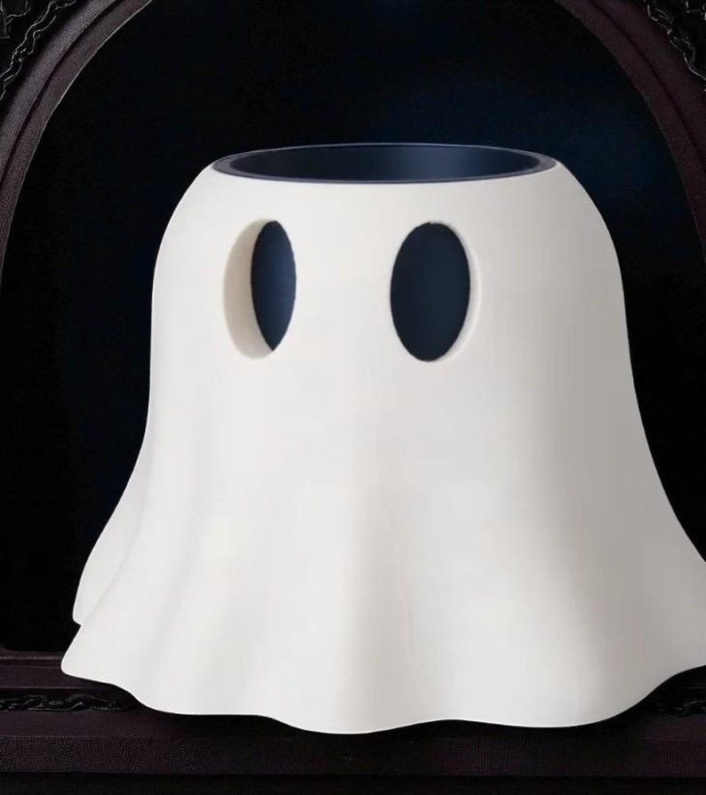 May include: A white ceramic ghost-shaped container with two oval eye-holes. The container has a wide, ruffled base and a dark interior. The design is simple and whimsical, perfect for Halloween or spooky decor.
