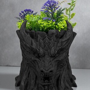 May include: A black, dragon-themed planter pot filled with artificial purple and green plants. The pot is sculpted with a detailed dragon face and textured bark-like appearance, creating a unique decorative piece.