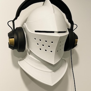 May include: A white, 3D-printed knight's helmet with a black headset. The helmet has a visor with small holes and is mounted on a wall. The headset has black earcups with gold accents.