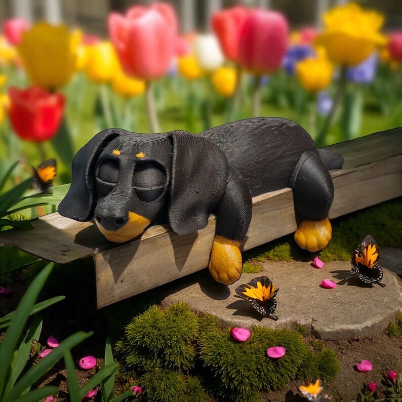 Pu&ograve; includere: Una scultura di cane bassotto nero e giallo appoggiata su una panchina di legno. Il cane ha gli occhi chiusi e dettagli gialli sulle zampe e sul muso. Farfalle e petali di fiori rosa sono sparsi intorno alla base della panchina, con un colorato giardino fiorito sullo sfondo.