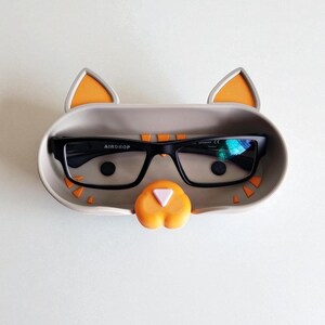 May include: A novelty glasses holder in the shape of a cat, in grey with orange details. The holder contains a pair of black-framed glasses. The glasses have the word "AIRDROP" printed on the frame.