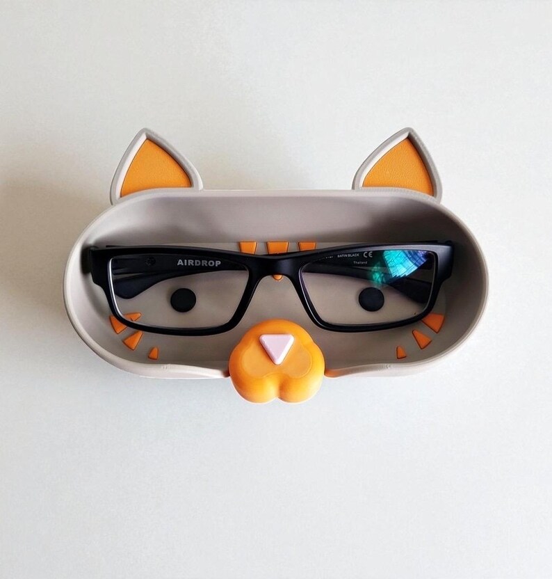 May include: A novelty glasses holder in the shape of a cat, in grey with orange details. The holder contains a pair of black-framed glasses. The glasses have the word "AIRDROP" printed on the frame.