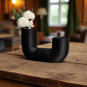 May include: A black, U-shaped vase with two openings, holding white flowers and green grass. The vase sits on a wooden table. The background is blurred, showing a room with a window and a lamp.