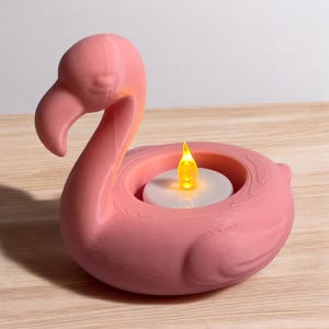 May include: Pink flamingo-shaped candle holder with a built-in space for a tea light. The flamingo has a curved neck and a detailed, textured surface. A yellow LED tea light is inside the holder, creating a warm glow. The candle holder is made of a solid material.