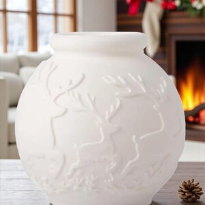 May include: A white ceramic vase with a round body and a slightly flared top. The vase features a raised relief design of reindeer and foliage. A small pinecone sits on the wooden surface next to the vase. The background includes a fireplace and window.