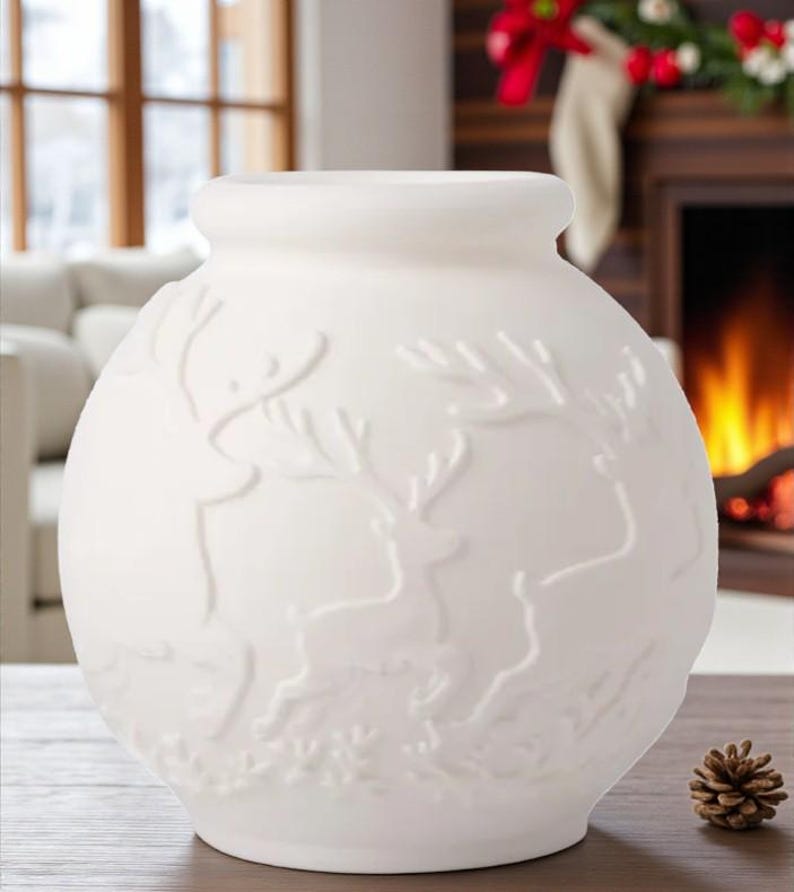May include: A white ceramic vase with a round body and a slightly flared top. The vase features a raised relief design of reindeer and foliage. A small pinecone sits on the wooden surface next to the vase. The background includes a fireplace and window.