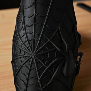 May include: A black, cylindrical vase featuring a detailed spiderweb design with a prominent spider accent. The vase is made of a matte material and sits on a wooden surface. The design is a 3D rendering.