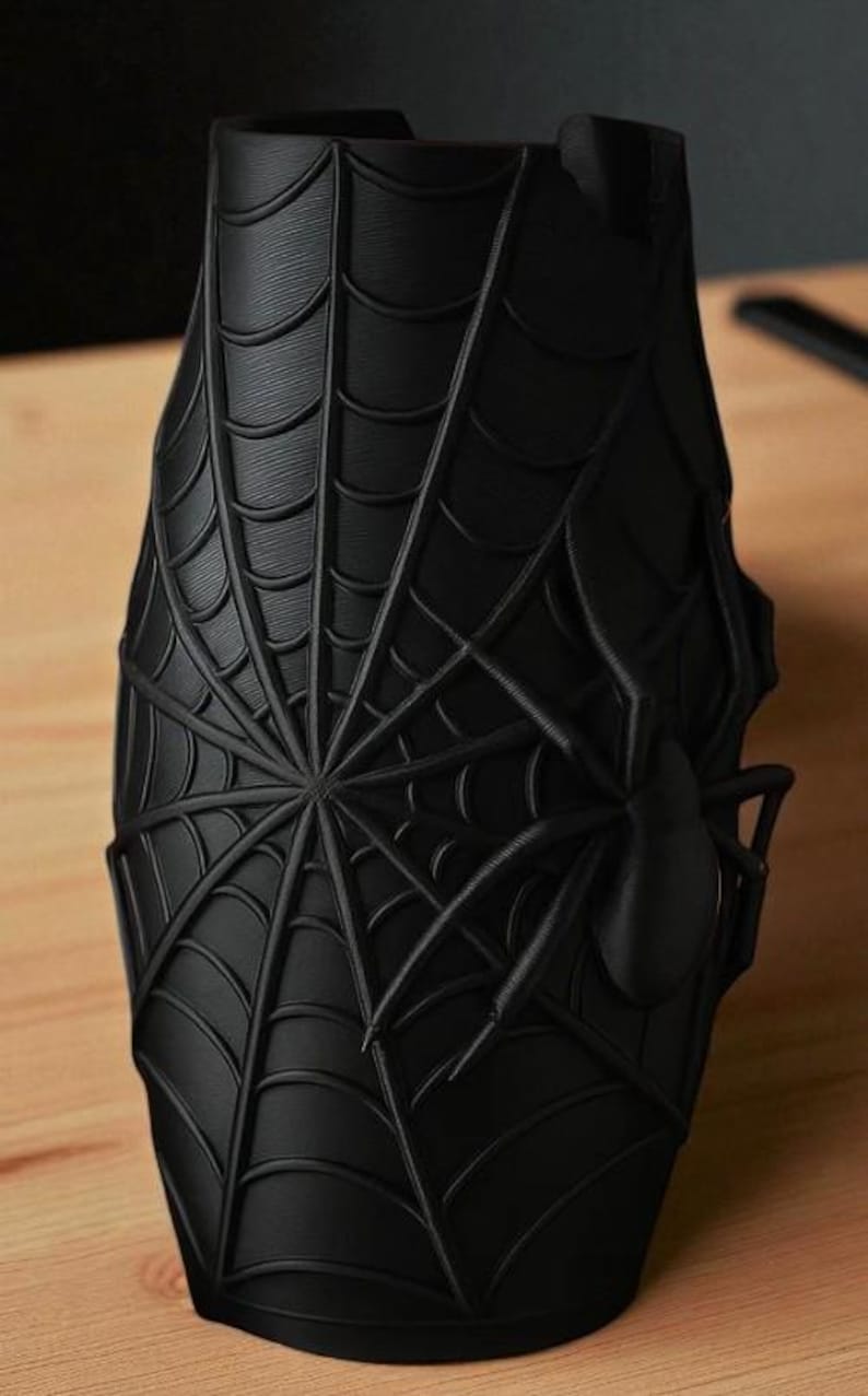May include: A black, cylindrical vase featuring a detailed spiderweb design with a prominent spider accent. The vase is made of a matte material and sits on a wooden surface. The design is a 3D rendering.