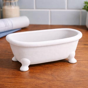 May include: A small, white ceramic bathtub with rounded edges and four small, rounded feet. The miniature tub is a decorative item, likely for display or as a container. It sits on a wooden surface.