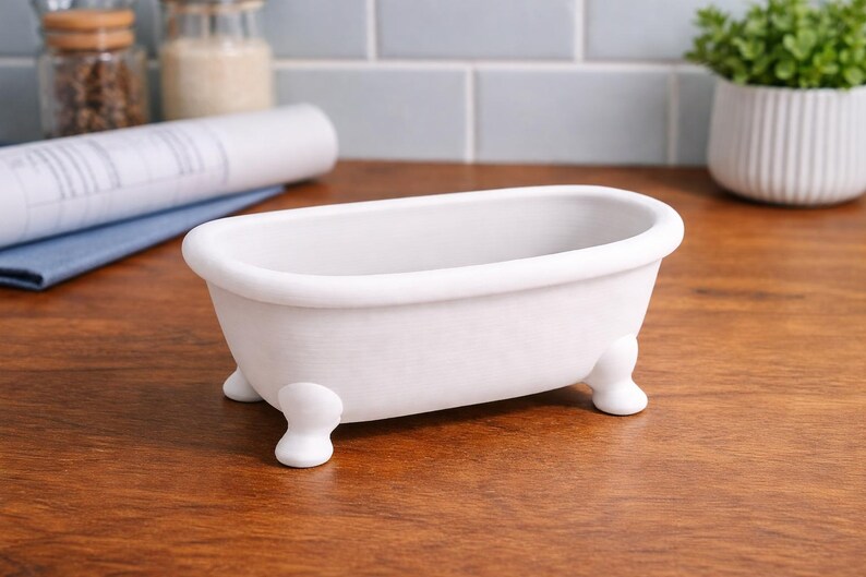 May include: A small, white ceramic bathtub with rounded edges and four small, rounded feet. The miniature tub is a decorative item, likely for display or as a container. It sits on a wooden surface.