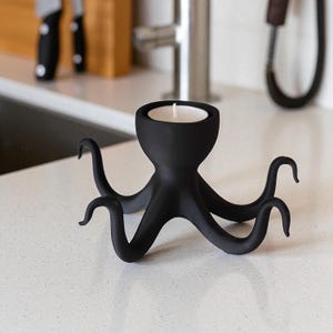 Op de afbeelding: Een zwarte kandelaar in de vorm van een octopus met een witte kaars. De houder heeft acht gebogen tentakels en een ronde beker voor de kaars. De kandelaar is gemaakt van matzwart materiaal en staat op een wit aanrecht.