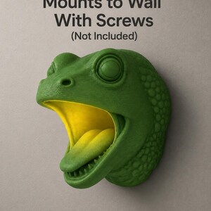 Op de afbeelding: Een groene dinosaurushoofdwandmontage met een gele binnenkant van de mond en tong. De kop is getextureerd en heeft een wijd open mond. De tekst "Mounts to Wall With Screws (Not Included)" staat bovenaan.
