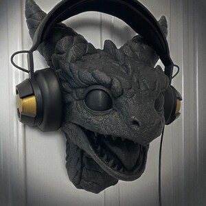 May include: A black dragon head wall mount with headphones. The dragon features detailed scales, horns, and an open mouth. The headphones are black with gold accents. The wall mount is designed to hold headphones.
