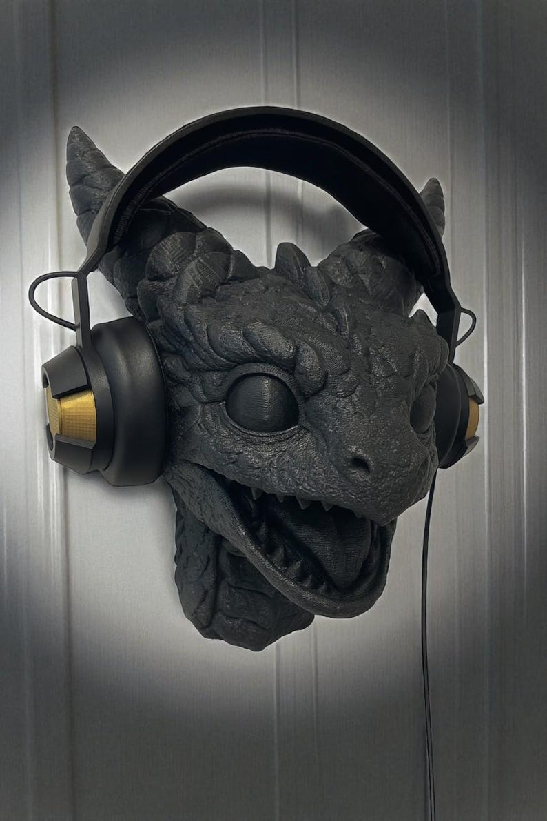 May include: A black dragon head wall mount with headphones. The dragon features detailed scales, horns, and an open mouth. The headphones are black with gold accents. The wall mount is designed to hold headphones.