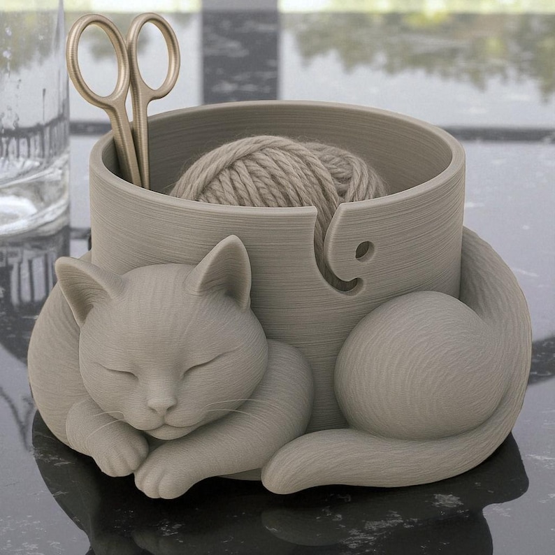 May include: A gray cat-shaped yarn bowl with a ball of yarn inside. The cat is curled up, with its eyes closed. A pair of gold scissors is placed in the bowl. The bowl has a hole for the yarn to feed through.