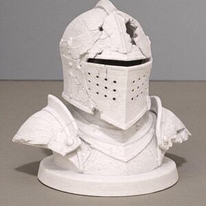 Puede incluir: Busto blanco de una armadura de caballero, que incluye un casco con visera y protectores de hombros. El casco tiene un aspecto da&ntilde;ado con grietas y un agujero. La armadura est&aacute; montada sobre una base circular.