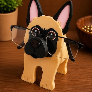 May include: A tan and black dog-shaped eyeglasses holder with a black nose and ears, holding a pair of black-framed glasses. The dog has pink inner ears and is made of wood.