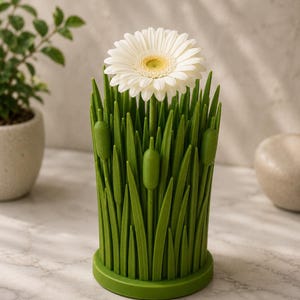 May include: A green, grass-like vase holds a white gerbera daisy with a yellow center. The vase has a cylindrical shape with a round base and is decorated with long, green blades of grass and three cattails.