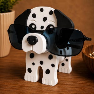 K&ouml;nnte beinhalten: Ein wei&szlig;-schwarzer Dalmatiner-Sonnenbrillenhalter. Der Halter hat schwarze Sonnenbrillen auf der Nase. Der Hund hat schwarze Ohren und schwarze Flecken auf seinem K&ouml;rper. Der Halter steht auf einer Holzoberfl&auml;che.